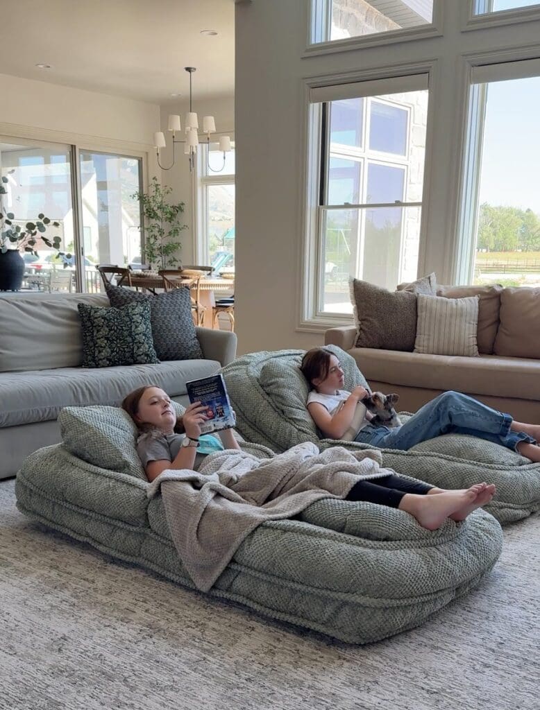 Two girls relaxing and reading books on side-by-side adjustable human dog beds in a sunlit living room.