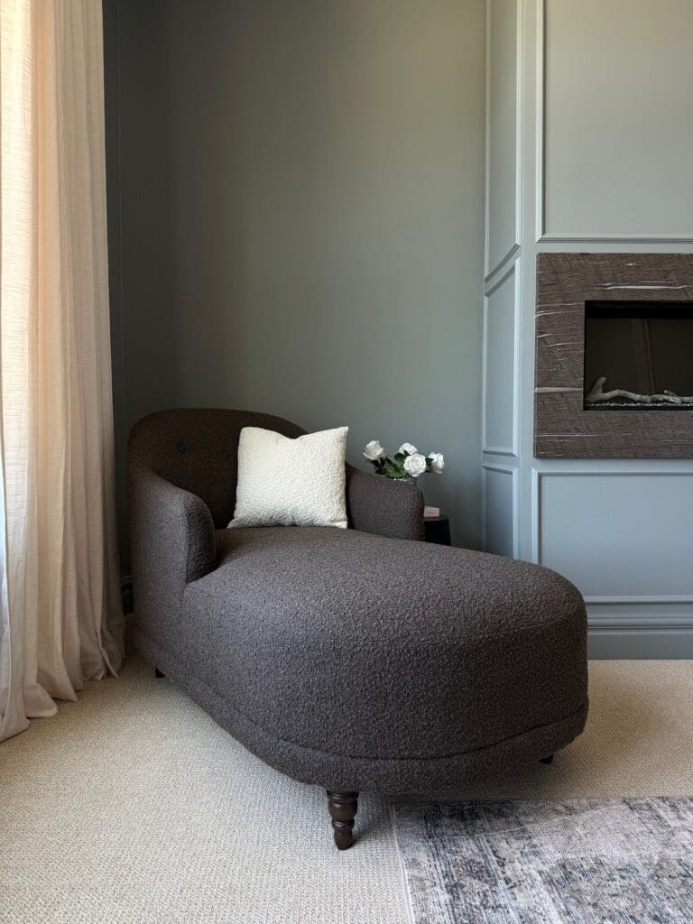 Cozy corner chaise lounge upholstered in textured dark fabric, next to fireplace and window.