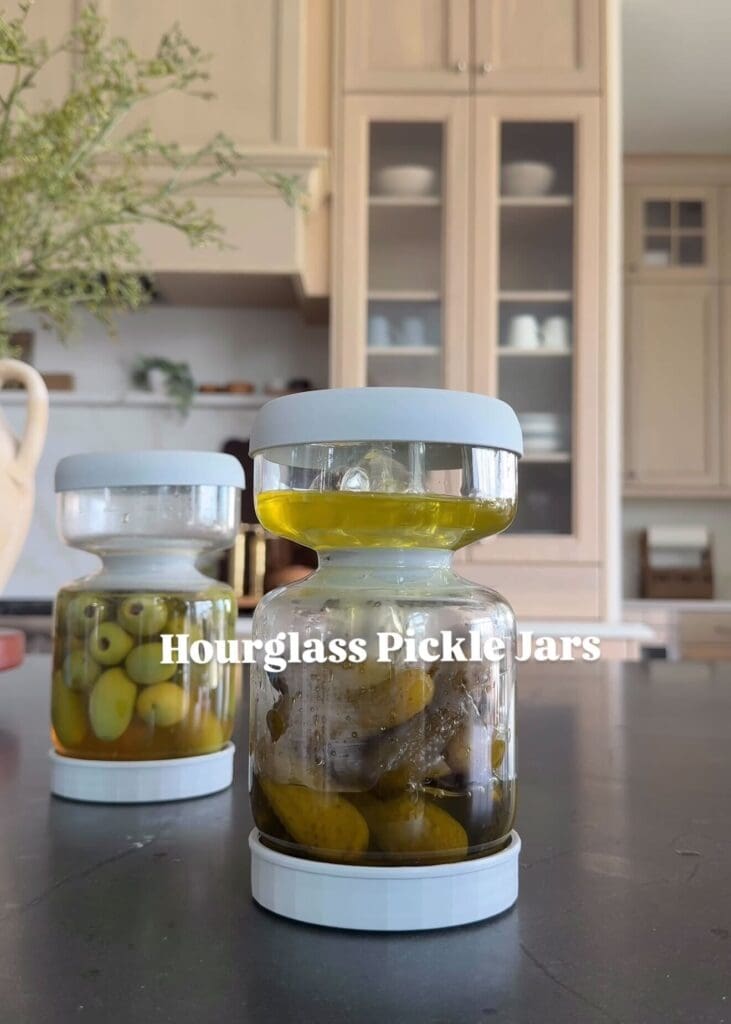Two hourglass-shaped pickle jars filled with olives and pickles on a dark countertop