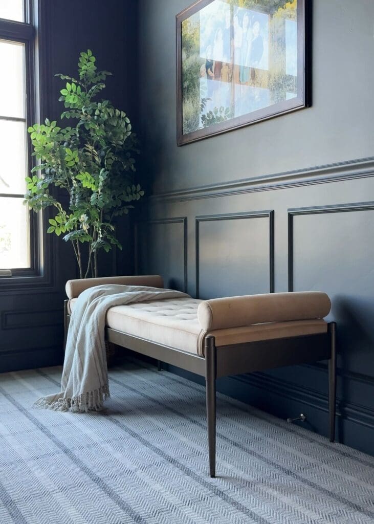 Full shot of velvet bolster bench styled with blanket in hallway with dark blue paneling and family photo above.