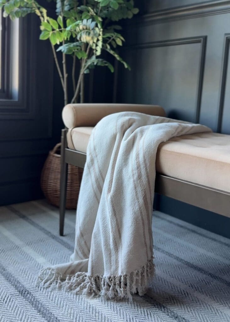 Beige velvet bolster bench styled under a window with a cozy fringed throw blanket draped across.