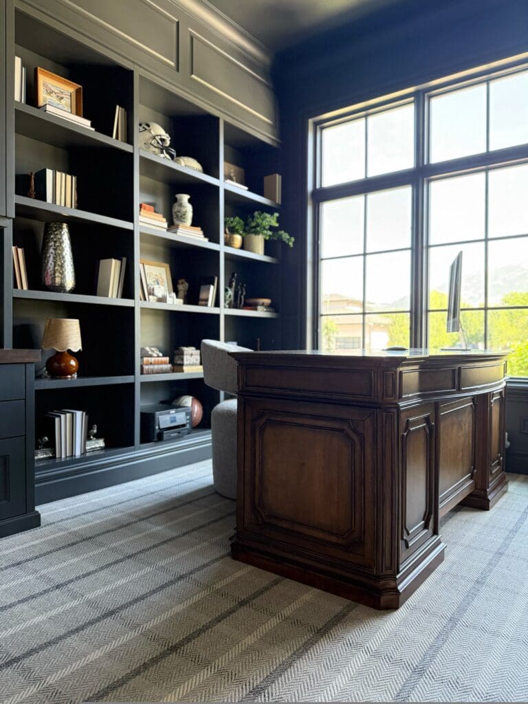 Dark charcoal built-in bookcases with styled shelves, a large wood desk, and striped neutral rug in a home office