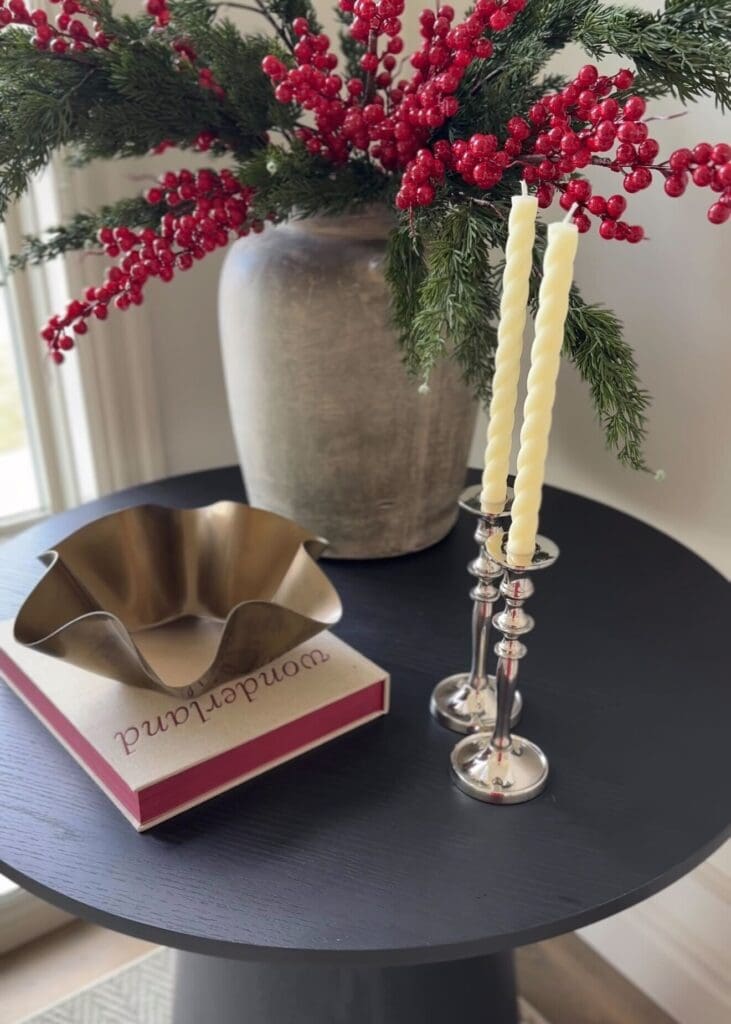Close-up of evergreen and berry arrangement in a stone vase on a black table with candles and a decorative bowl.