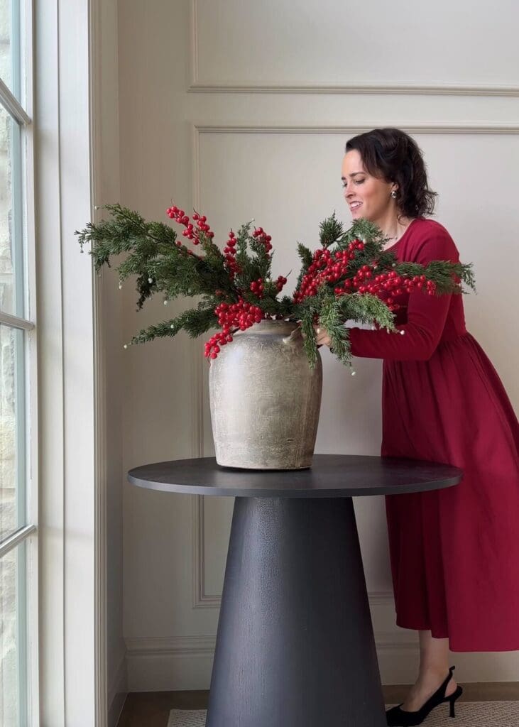 Andrea in a red dress styling a stone vase filled with evergreen and red berry stems on a black round pedestal table in a bright entryway.