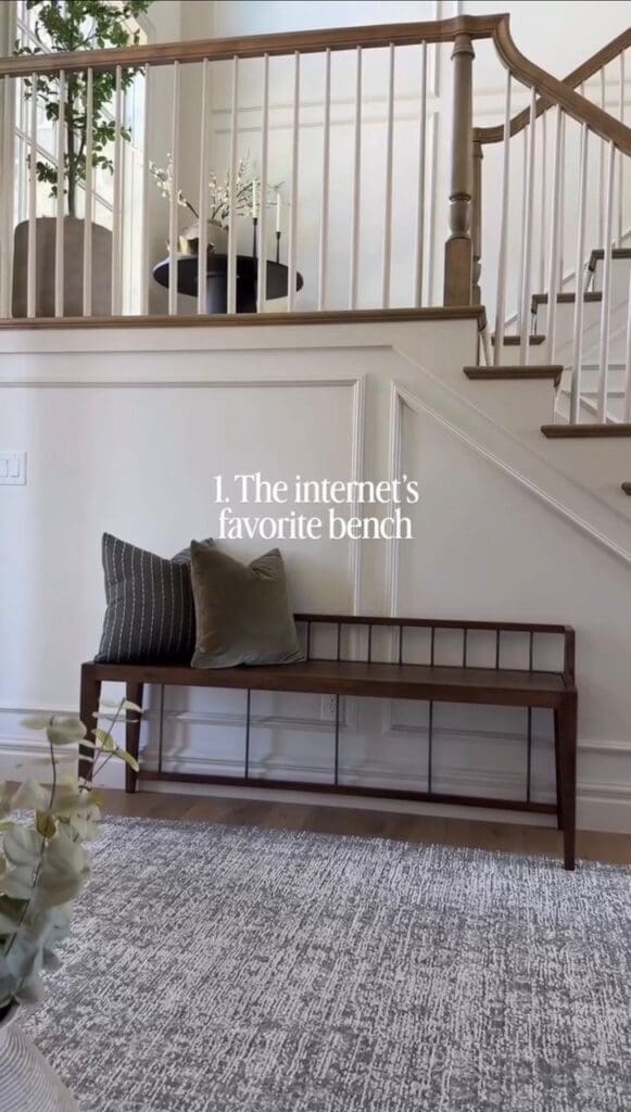 Wood entryway bench styled under a staircase with neutral pillows and classic wall molding.
