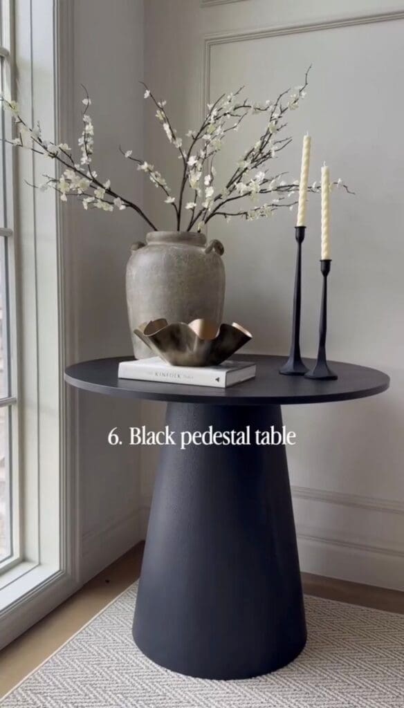 Black pedestal accent table styled with neutral decor and candles near a window.

